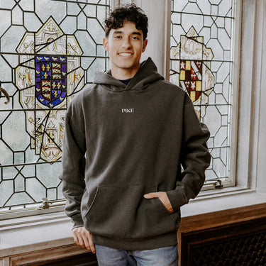 College-aged man wearing the Pi Kappa Alpha embroidered black hoodie, standing in front of stained glass windows with fraternity-style crest designs.