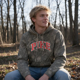 College-aged male wearing the fraternity Deadwood Camo Hoodie with red and white Pi Kappa Alpha lettering, seated outdoors in a wooded setting.