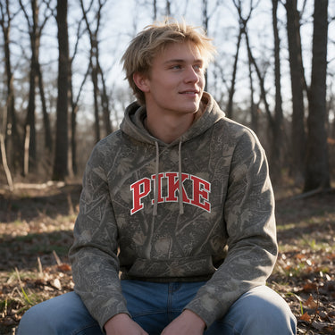College-aged male wearing the fraternity Deadwood Camo Hoodie with red and white Pi Kappa Alpha lettering, seated outdoors in a wooded setting.
