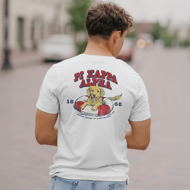 Pi Kappa Alpha  fraternity member in a limited edition  white short sleeve tee  featuring a golden retriever puppy playing in an pool floatie  with the fraternity name written above and the founding year.