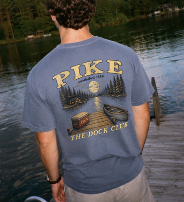 Young man wearing a blue graphic tee standing on a lakeside dock, showing the back design