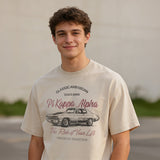 Young man wearing a vintage-style beige graphic T-shirt featuring a retro muscle car illustration and cursive text, standing outdoors with a neutral blurred background.