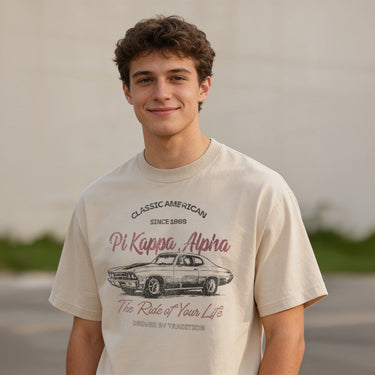 Young man wearing a vintage-style beige graphic T-shirt featuring a retro muscle car illustration and cursive text, standing outdoors with a neutral blurred background.