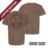 Brown garment-dyed short-sleeve tee featuring a vintage-inspired graphic: a weathered oval frames a mallard in mid-flight with burnt orange and mossy green-tipped wings above rippling water and cattails; distressed Pi Kappa Alpha arcs overhead and PIKE letters bold and rugged on the front.